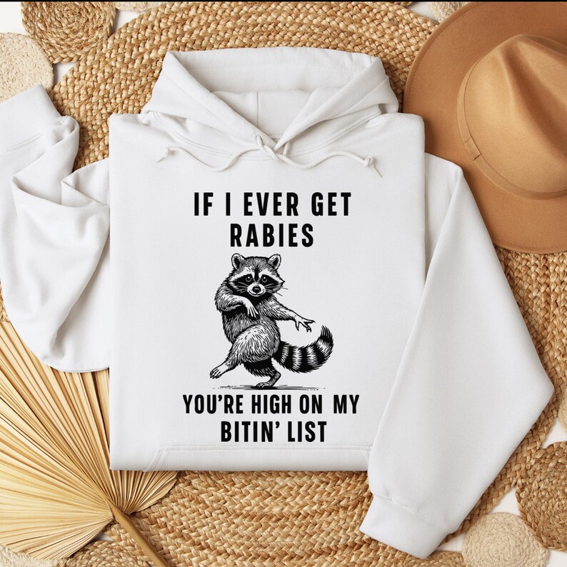 Op de afbeelding: Witte hoodie met een wasbeer-afbeelding en de tekst "IF I EVER GET RABIES YOU'RE HIGH ON MY BITIN' LIST". De wasbeer is afgebeeld in zwart en wit. De hoodie heeft een capuchon met trekkoord.