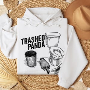 May include: White hoodie with a black and white illustration of a raccoon, a toilet, and a bin. The text "Trashed Panda" is printed above the graphic. The hoodie has a drawstring hood and long sleeves.