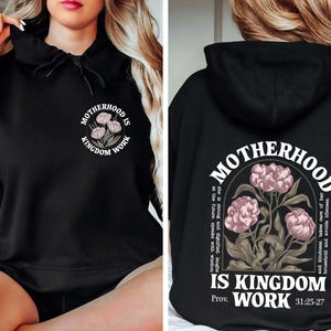 May include: Black hoodie with the text "MOTHERHOOD IS KINGDOM WORK" and a floral design. The design is on the front and back of the hoodie. The back of the hoodie also includes a Bible verse.