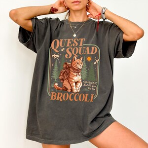 May include: A dark gray oversized t-shirt with a vintage-style graphic of a cat wearing a backpack and the text "QUEST SQUAD BROCCOLI". The graphic also includes the text "No one wants it, but it's good for you".