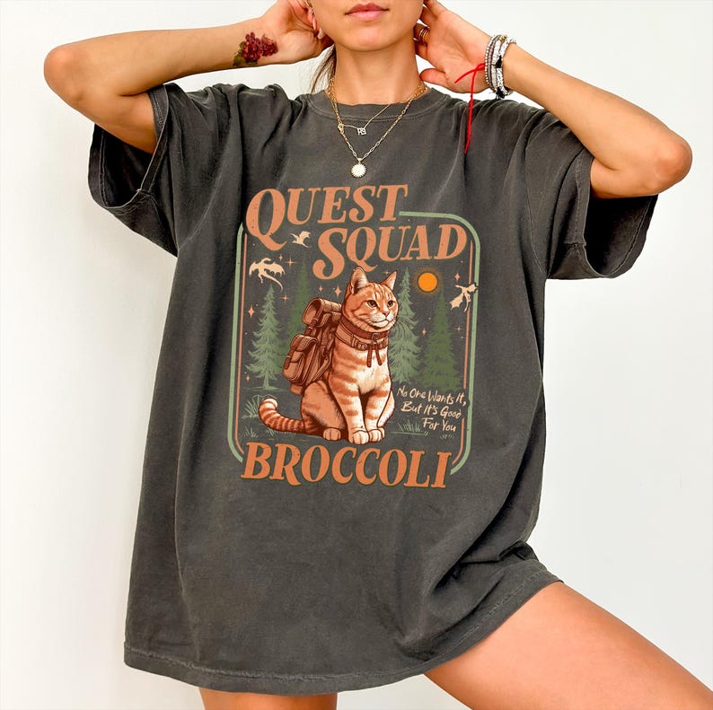 May include: A dark gray oversized t-shirt with a vintage-style graphic of a cat wearing a backpack and the text "QUEST SQUAD BROCCOLI". The graphic also includes the text "No one wants it, but it's good for you".