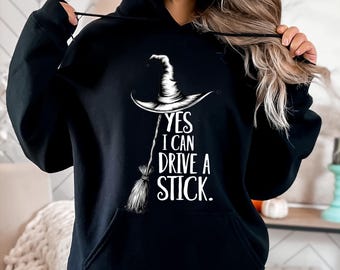 May include: Black hooded sweatshirt with a white graphic of a witch's hat and broom. The text reads "YES I CAN DRIVE A STICK." The sweatshirt has a front pocket and a drawstring hood.