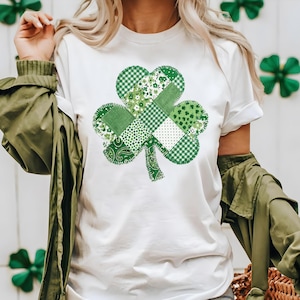 May include: White t-shirt featuring a large, patchwork shamrock design in various shades of green. The shamrock is composed of different patterns, including gingham, floral, and paisley. The shirt has short sleeves and a crew neck.