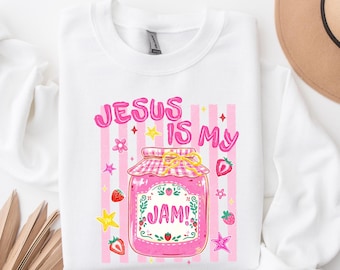 Puede incluir: Una sudadera blanca de cuello redondo con un gráfico de un tarro de mermelada rosa con tapa a cuadros. El texto "JESUS IS MY JAM!" está rodeado de ilustraciones de fresas, estrellas amarillas y flores rosas sobre un fondo de rayas rosas.