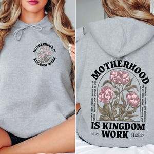 May include: A light gray hoodie with the text "MOTHERHOOD IS KINGDOM WORK" and a floral design. The back of the hoodie features the same text and design, along with additional text. The hoodie has a drawstring hood.