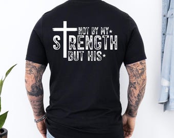 May include: Black t-shirt with a white cross and the text "NOT BY MY STRENGTH BUT HIS" in a bold, stylized font. The design includes star accents. The shirt is a classic crew neck.