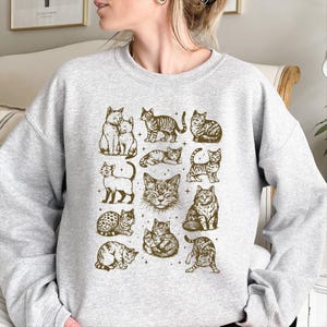 May include: A light gray sweatshirt featuring a repeating pattern of various cat illustrations in a sepia tone. The cats are depicted in different poses and styles, creating a charming and whimsical design. The sweatshirt is a comfortable and casual garment.