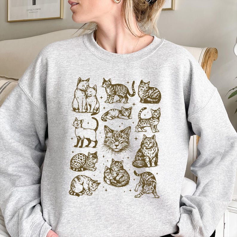 May include: A light gray sweatshirt featuring a repeating pattern of various cat illustrations in a sepia tone. The cats are depicted in different poses and styles, creating a charming and whimsical design. The sweatshirt is a comfortable and casual garment.