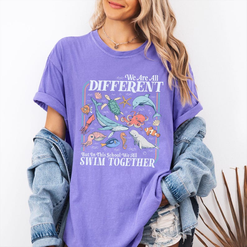 May include: A purple t-shirt with the text "We Are All Different But In This School We All Swim Together" and various sea creatures. The graphic includes a whale, octopus, dolphin, and other marine life.