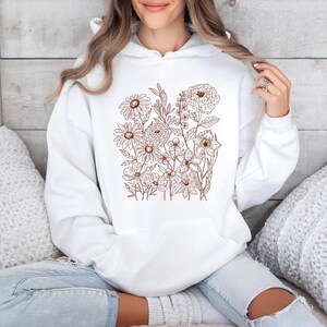 May include: White hoodie featuring a brown floral design. The design includes various flowers and foliage, creating a botanical illustration. The hoodie has a front pocket and a drawstring hood, offering a casual and comfortable style.