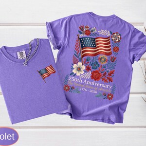May include: Two violet t-shirts. One has a small American flag embroidered on the chest. The other has a large American flag design on the back, surrounded by flowers, with the text "250th Anniversary The United States of America 1776-2026".