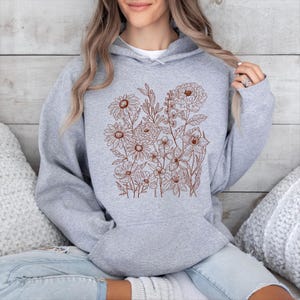 May include: A light gray hoodie featuring a brown floral design. The design includes various flowers and foliage, creating a botanical theme. The hoodie has a drawstring hood and a kangaroo pocket.