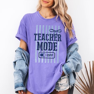 May include: A purple t-shirt with the text "TEACHER MODE OFF" in navy blue and light blue. The shirt has vertical light blue stripes and a bow graphic above the text. The model is wearing a denim jacket.