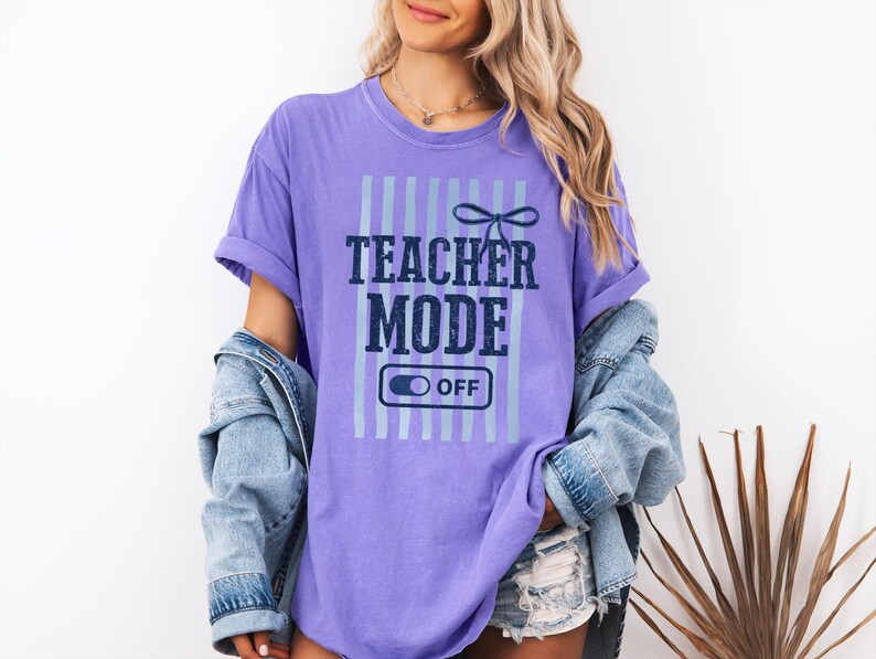 May include: A purple t-shirt with the text "TEACHER MODE OFF" in navy blue and light blue. The shirt has vertical light blue stripes and a bow graphic above the text. The model is wearing a denim jacket.