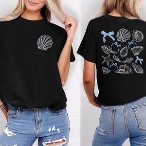 Puede incluir: Camiseta negra con un dise&ntilde;o de concha en la parte delantera y una colecci&oacute;n de ilustraciones de conchas marinas con lazos azules en la parte trasera. El dise&ntilde;o incluye varios tipos de conchas y una estrella de mar. La camiseta es de estilo casual y c&oacute;modo.