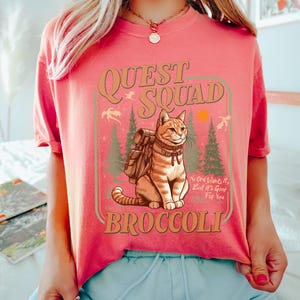 May include: A pink t-shirt with a graphic of a cat wearing a backpack and the text "Quest Squad Broccoli" with the tagline "No one wants it, but it's good for you".