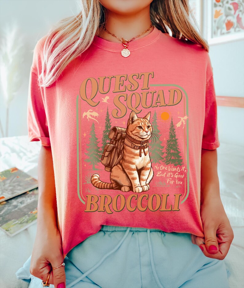May include: A pink t-shirt with a graphic of a cat wearing a backpack and the text "Quest Squad Broccoli" with the tagline "No one wants it, but it's good for you".