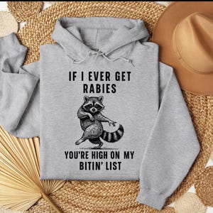 Op de afbeelding: Een lichtgrijze hoodie met de tekst "IF I EVER GET RABIES YOU'RE HIGH ON MY BITIN' LIST" en een dansende wasbeer-afbeelding in zwart en wit. De hoodie heeft een capuchon met trekkoord en een kangoeroezak.