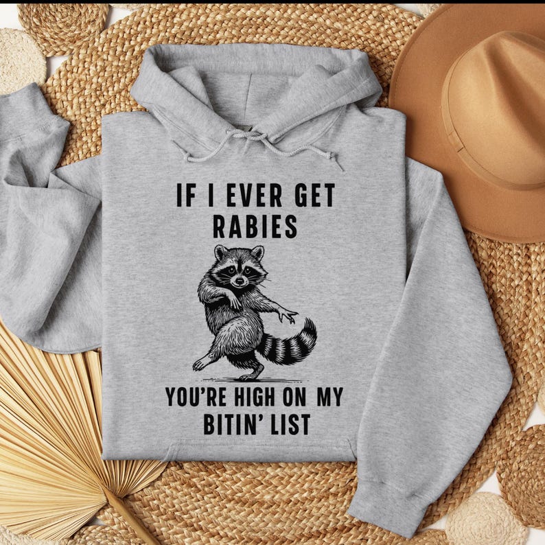 Op de afbeelding: Een lichtgrijze hoodie met de tekst "IF I EVER GET RABIES YOU'RE HIGH ON MY BITIN' LIST" en een dansende wasbeer-afbeelding in zwart en wit. De hoodie heeft een capuchon met trekkoord en een kangoeroezak.