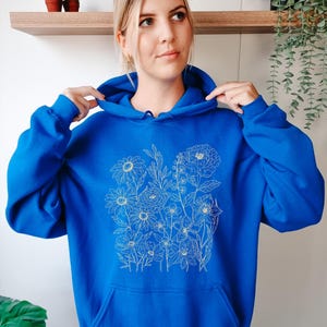 May include: A royal blue hoodie featuring a floral design in a delicate, off-white line drawing. The hoodie has a front pocket and a drawstring hood. The floral design includes daisies, peonies, and other flowers.