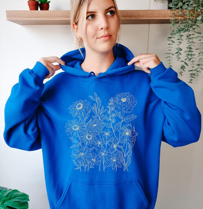 May include: A royal blue hoodie featuring a floral design in a delicate, off-white line drawing. The hoodie has a front pocket and a drawstring hood. The floral design includes daisies, peonies, and other flowers.
