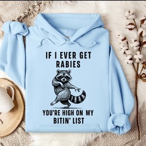 Op de afbeelding: Een lichtblauwe hoodie met de tekst "IF I EVER GET RABIES YOU'RE HIGH ON MY BITIN' LIST" en een zwart-wit wasbeer-afbeelding. De wasbeer wordt afgebeeld in een danshouding. De hoodie heeft een trekkoord en een kangoeroezak.