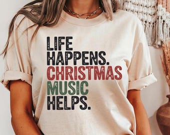 May include: A beige t-shirt with the text "LIFE HAPPENS. CHRISTMAS MUSIC HELPS." in black, red, and green lettering. The shirt has short sleeves and a crew neck.