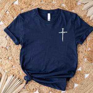 May include: A navy blue t-shirt with a white cross design featuring nails. The cross is centered on the shirt.