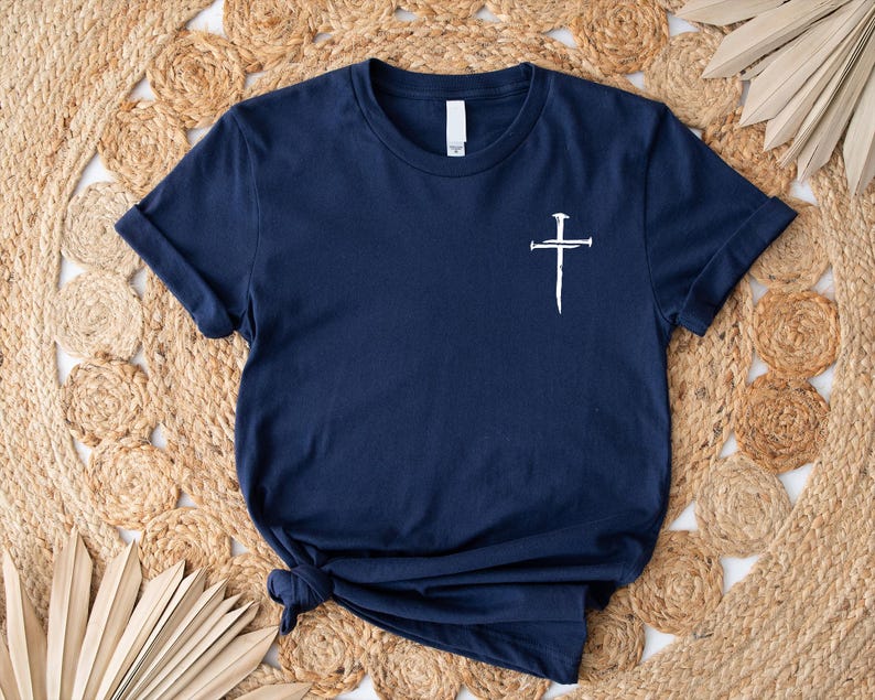 May include: A navy blue t-shirt with a white cross design featuring nails. The cross is centered on the shirt.
