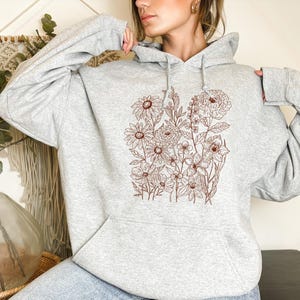 May include: A light gray hoodie featuring a brown floral design. The design includes various flowers and foliage, creating a botanical illustration. The hoodie has a front pocket and a drawstring hood.