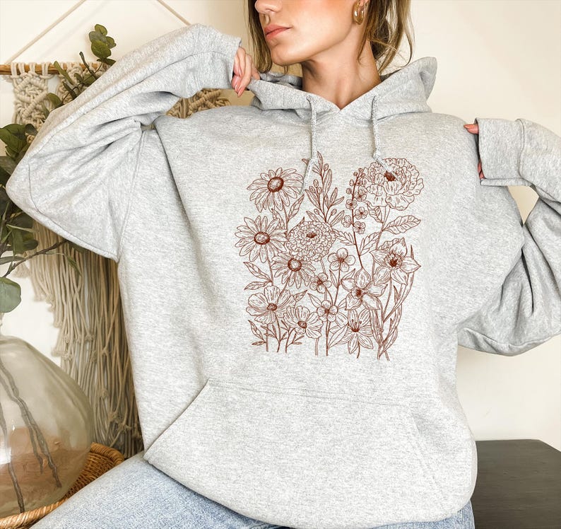 May include: A light gray hoodie featuring a brown floral design. The design includes various flowers and foliage, creating a botanical illustration. The hoodie has a front pocket and a drawstring hood.