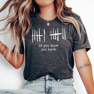 May include: Dark gray t-shirt with white text that reads "if you know you know" below a series of vertical lines. The shirt is a crew neck and is worn with blue jeans. The model is wearing bracelets and a necklace.