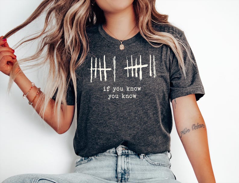 May include: Dark gray t-shirt with white text that reads "if you know you know" below a series of vertical lines. The shirt is a crew neck and is worn with blue jeans. The model is wearing bracelets and a necklace.