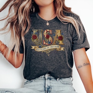 May include: Dark grey t-shirt with the word "USA" in gold, floral-decorated letters. The shirt features red roses and a banner that reads "250 Years of Freedom". The shirt is worn by a person.