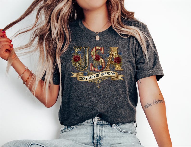 May include: Dark grey t-shirt with the word "USA" in gold, floral-decorated letters. The shirt features red roses and a banner that reads "250 Years of Freedom". The shirt is worn by a person.