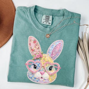 May include: A teal Comfort Colors t-shirt featuring a colorful bunny design. The bunny has floral ears, heart-shaped glasses, and a floral crown. A silver necklace with a small pendant is draped across the shirt.