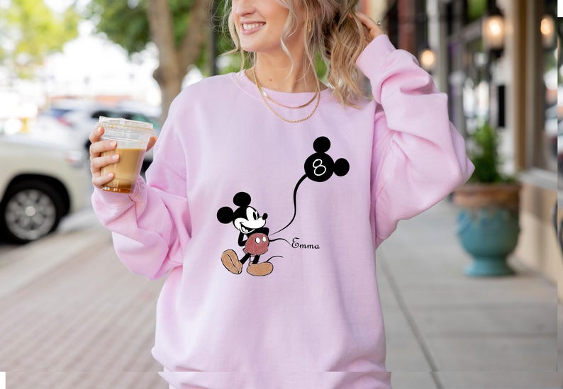 Puede incluir: Sudadera rosa con un gr&aacute;fico de Mickey Mouse en blanco y negro sosteniendo un globo con el n&uacute;mero 8. El texto "Emma" est&aacute; impreso debajo del gr&aacute;fico.