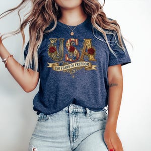 May include: A navy blue t-shirt featuring the word "USA" in an ornate design, embellished with red roses and gold detailing. The banner below reads "250 YEARS OF FREEDOM". The shirt is worn with light blue denim jeans.
