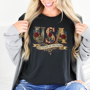 May include: Black t-shirt with a decorative "USA" design featuring floral accents and the text "250 YEARS OF FREEDOM" on a banner. The letters are gold with red, white, and blue details.