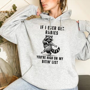Op de afbeelding: Een lichtgrijze hoodie met een voorzak en een trekkoord. De voorkant heeft een zwart-wit illustratie van een wasbeer en de tekst "IF I EVER GET RABIES YOU'RE HIGH ON MY BITIN' LIST."