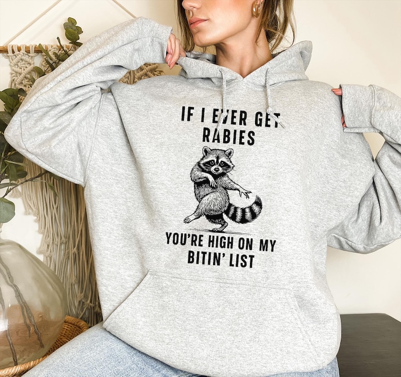 Op de afbeelding: Een lichtgrijze hoodie met een voorzak en een trekkoord. De voorkant heeft een zwart-wit illustratie van een wasbeer en de tekst "IF I EVER GET RABIES YOU'RE HIGH ON MY BITIN' LIST."