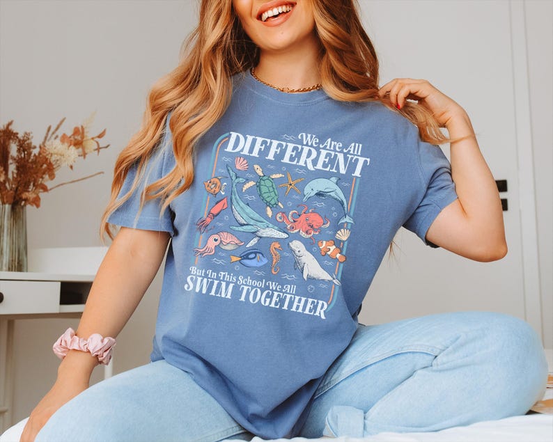 May include: Blue t-shirt with a colorful graphic of various sea creatures, including a whale, octopus, and fish. The text on the shirt reads "We Are All DIFFERENT But In This School We All SWIM TOGETHER."