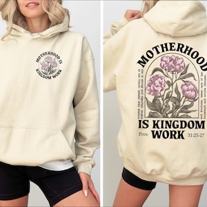 May include: Cream-colored hoodie with a front pocket and hood. The front features a circular design with the text "MOTHERHOOD IS KINGDOM WORK" and a floral illustration. The back has the same text and floral design, with additional text and scripture reference.