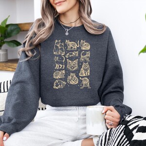 May include: Dark gray sweatshirt featuring a gold cat illustration. The design includes various cat poses and faces. The sweatshirt has long sleeves and a crew neck. The person in the image is holding a white mug.