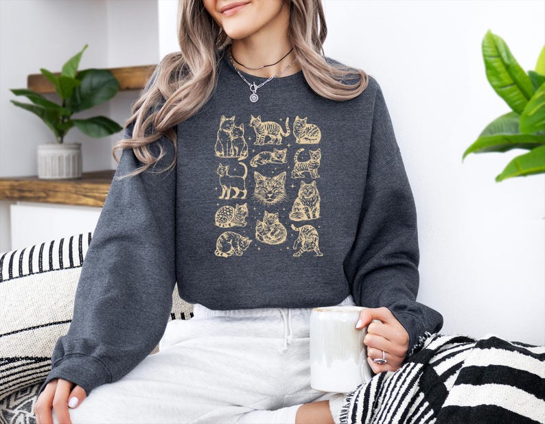 May include: Dark gray sweatshirt featuring a gold cat illustration. The design includes various cat poses and faces. The sweatshirt has long sleeves and a crew neck. The person in the image is holding a white mug.