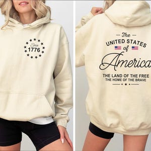 Pu&ograve; includere: Felpa con cappuccio color crema con tasca frontale e cappuccio. La parte anteriore presenta un design circolare con la scritta "Since 1776". Il retro mostra "The United States of America" con bandiere e le frasi "The Land of the Free" e "The Home of the Brave."