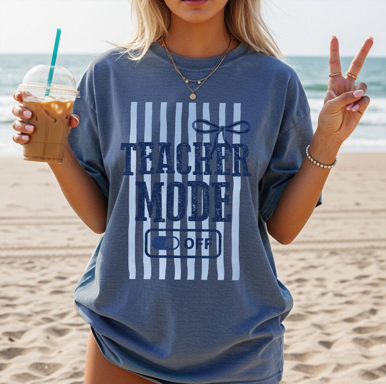 May include: A blue-grey t-shirt with the text "TEACHER MODE OFF" in navy blue. The text is set against a background of white vertical stripes. The shirt has a relaxed fit and short sleeves.