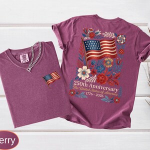 May include: Two berry-colored t-shirts. One has a small American flag patch, the other has a large American flag design with floral accents and text that reads "250th Anniversary The United States of America 1776-2026."