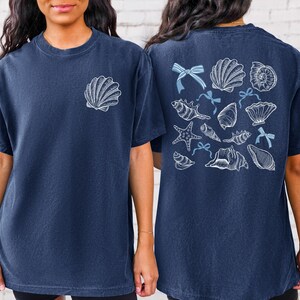 Puede incluir: Camiseta azul marino con un gr&aacute;fico de concha en la parte delantera y una colecci&oacute;n de conchas marinas, estrellas de mar y lazos en la parte trasera. El dise&ntilde;o es en blanco y azul claro. La camiseta est&aacute; hecha de un material suave y c&oacute;modo.