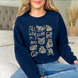 May include: Navy blue sweatshirt featuring a beige illustration of various cat poses. The design includes cats sitting, standing, and lying down. The sweatshirt is made of a soft material, perfect for casual wear.